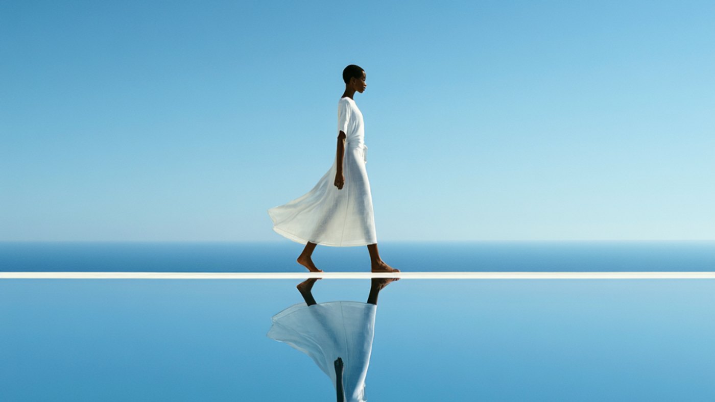 A woman in a white dress walking a mirrored ledge above a still blue sea